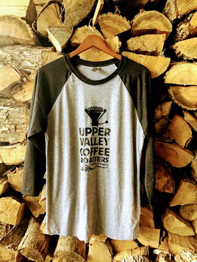 A grey and black 3/4 sleeve raglan tee with UV Coffee Roasters logo is hung in front of a stack of firewood.